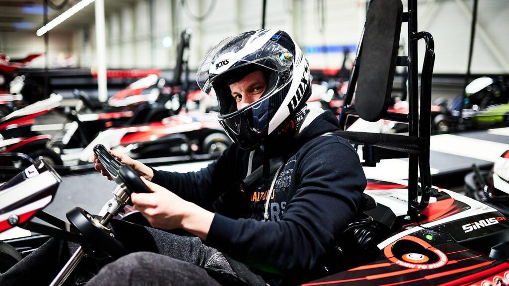 Ein Mann sitzt in einem Go-Kart und schaut direkt in die Kamera. Er trägt einen schwarzen Kapuzenpullover und einen weißen Helm mit Visier, der geöffnet ist. Im Hintergrund sind weitere Go-Karts auf der Strecke zu erkennen.