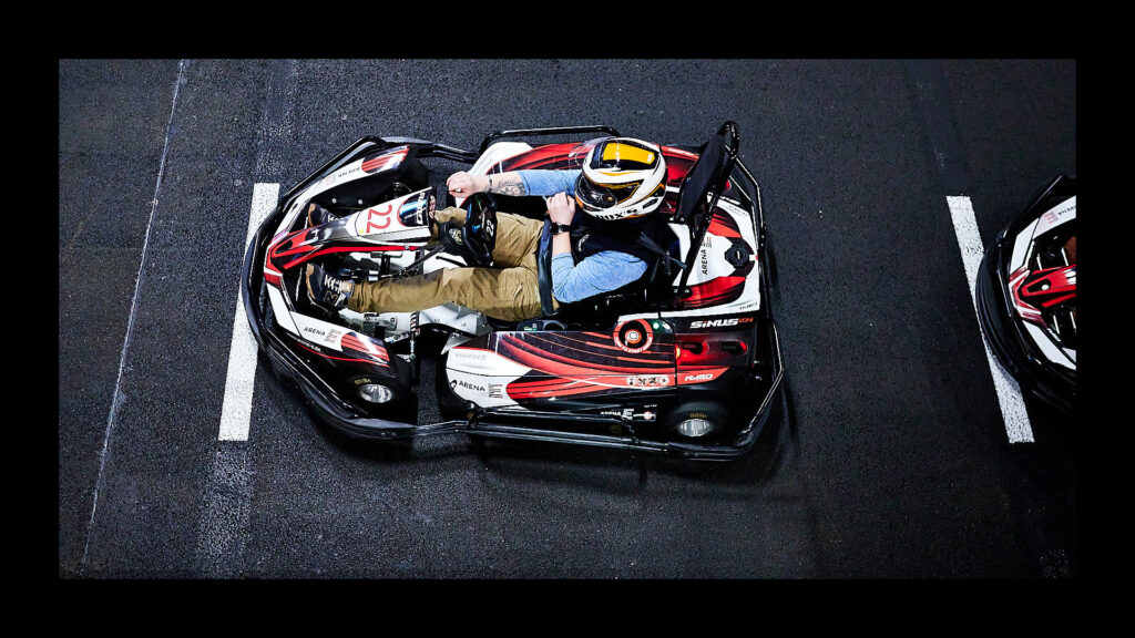 Eine Nahaufnahme aus der Vogelperspektive eines Rennfahrers, der einen schwarzen Go-Kart steuert. Er trägt einen Helm und hat die Hände am Lenkrad. Der Go-Kart ist weiß, rot und schwarz und auf dem Boden sind weiße Linien sichtbar.