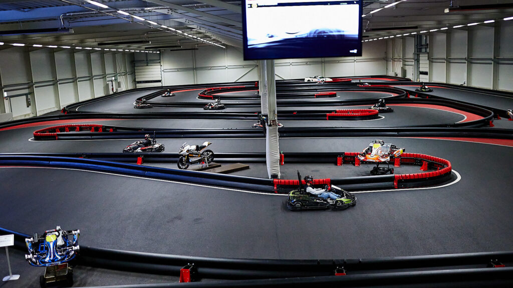 Ein Rennfahrer in einem schwarzen Go-Kart fährt eine Kurve auf einer beleuchteten Indoor-Go-Kart-Strecke. Im Hintergrund sind weitere Go-Karts, eine Zuschauertribüne und ein großer Bildschirm zu sehen.