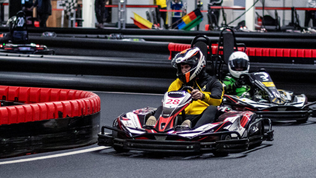 Ein Kart-Fahrer in einem schwarzen Rennanzug und Helm steuert sein Go-Kart auf der Strecke. Im Hintergrund sind weitere Go-Karts und die Zuschauergalerie zu sehen.