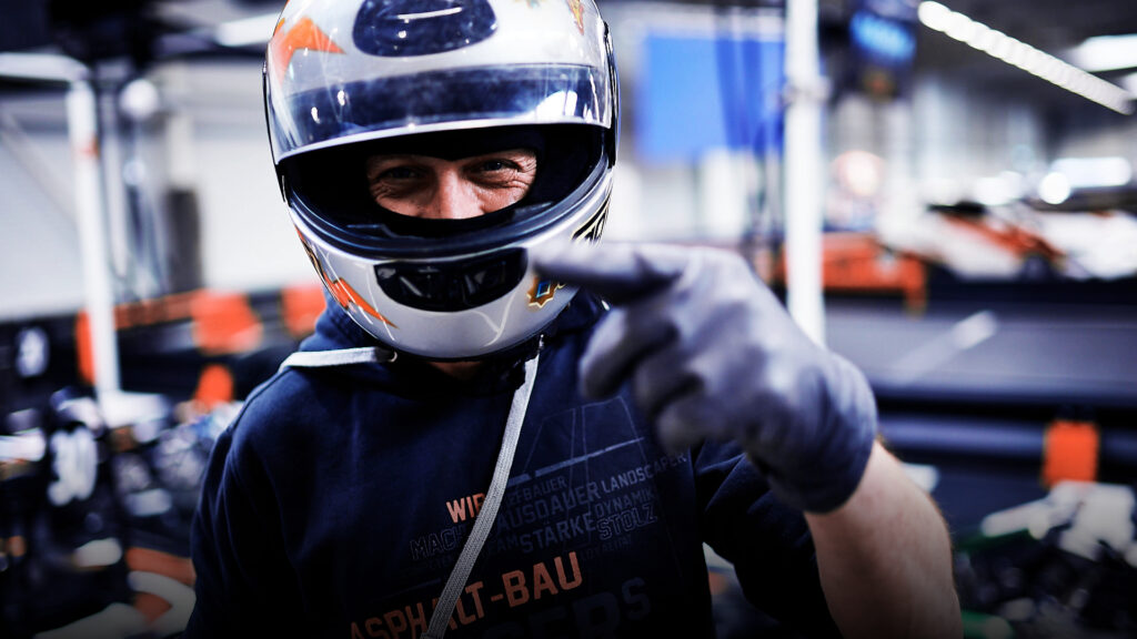 Nahaufnahme eines lächelnden Mannes mit Motorsporthelm und Handschuhen, der auf der Indoor-Kartbahn auf den Betrachter zeigt.