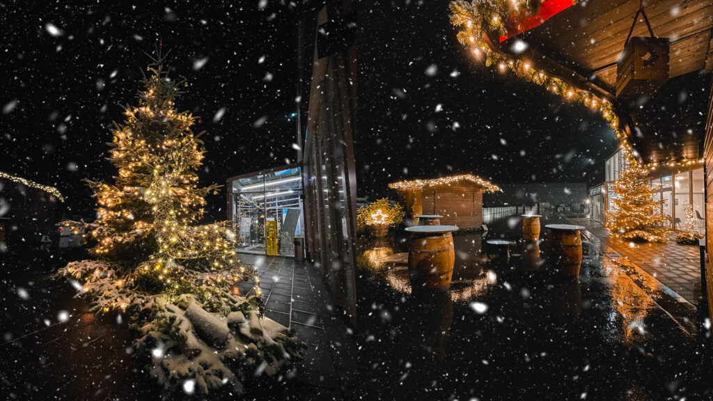 Winterliche Aufnahme eines Weihnachtsmarktes bei Nacht und Schneefall. Im Zentrum steht ein beleuchteter Tannenbaum, daneben rustikale Holzbuden und Stehtische aus Weinfässern. Die Szene ist festlich mit warmen Lichterketten geschmückt und vermittelt eine gemütliche Atmosphäre. Im Hintergrund ist die Arena E zu sehen