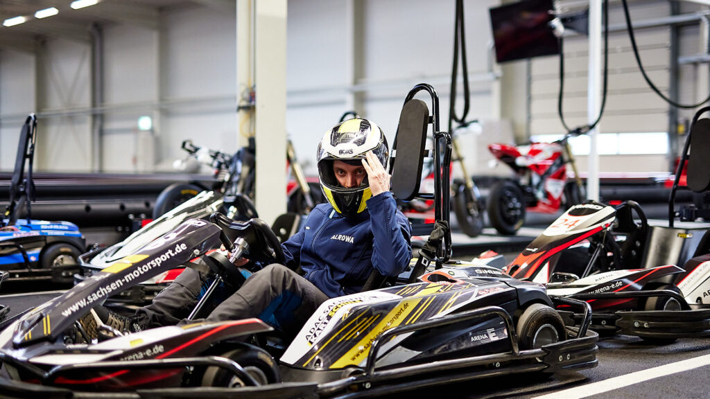Männliche person mit helm sitzend in einem kart, dahinter weitere Karts in der Indoor-Halle.